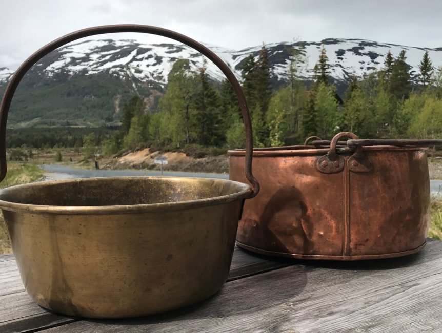 To gamle gryter på trebenk med snødekte fjell og skog i bakgrunnen.