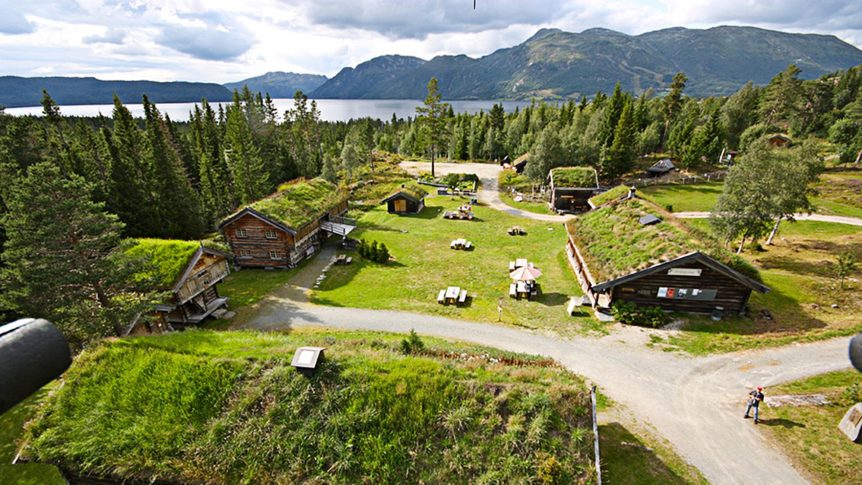 Tradisjonelle norske hus med torvtak omgitt av skog, fjell og innsjø i naturskjønn dal.
