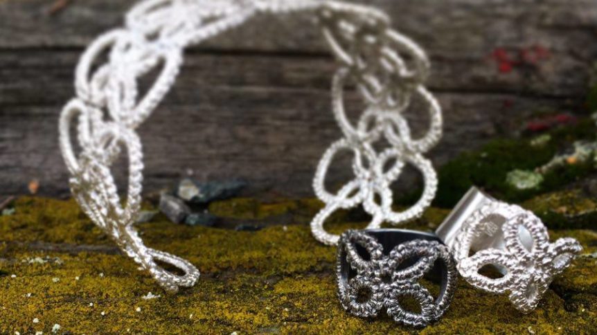 Håndlagde sølvsmykker, et armbånd, og to ringer med intrikate design mot en naturlig bakgrunn av mose og tre.