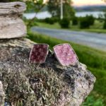 Rosa mansjettknapper med firkantet design på stein, omgitt av naturlig landskap i bakgrunnen.