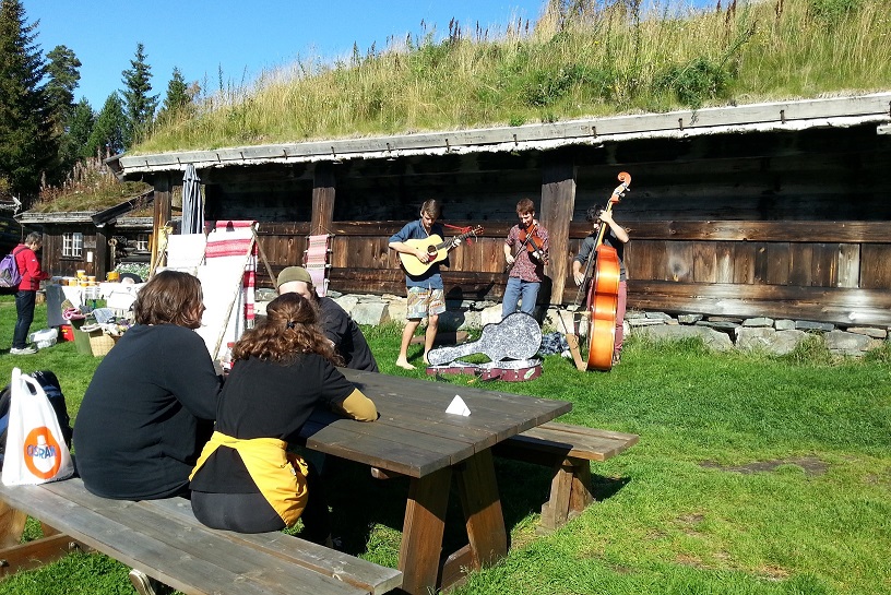 Gruppe lytter til levende musikk utendørs ved en tømmerhytte på en solrik dag i Norge.