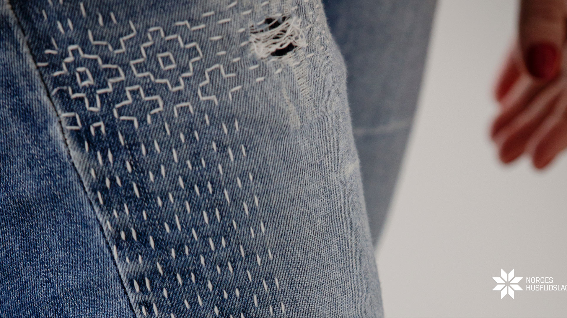 Detalj av blå jeans med dekorativt broderi, nærbilde. Hånd med rød neglelakk i bakgrunnen.