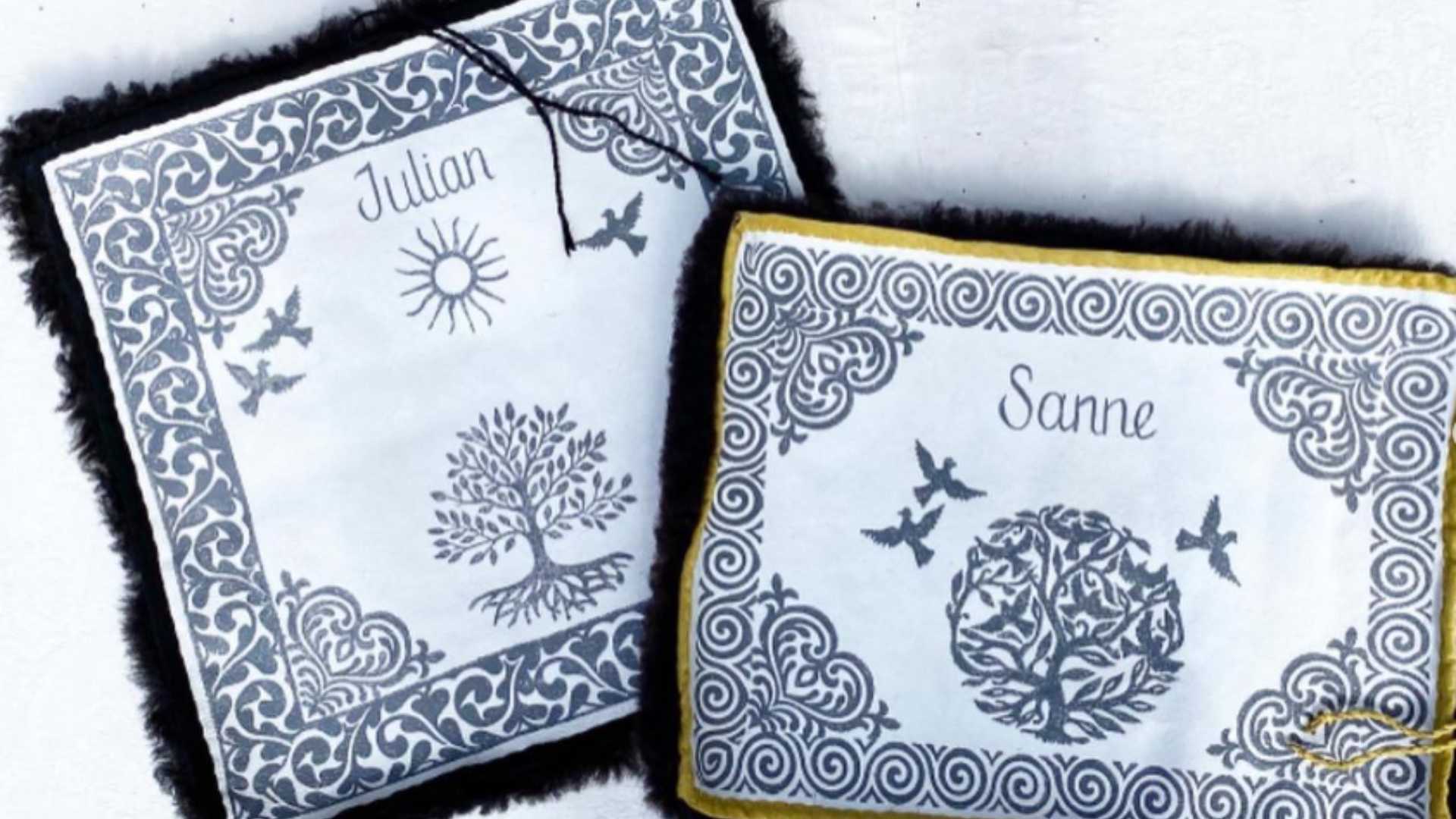To broderte puter med navnene Julian og Sanne, dekorert med mønstre av sol, trær og fugler.