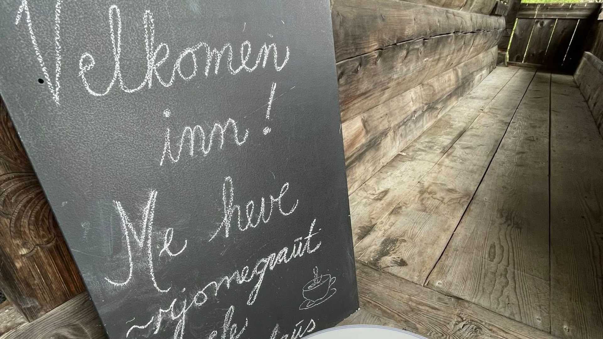 Tavle med teksten Velkomen inn! Me heve rjomegraut ved en trevegg i en koselig, rustikk setting.