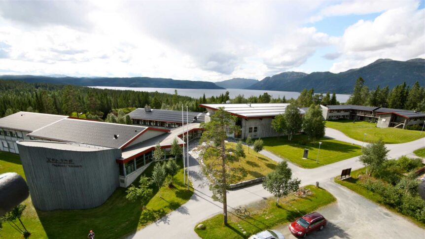 Utsikt over skolebygg omgitt av skog og fjord i Norge, med fjell i bakgrunnen under en overskyet himmel.