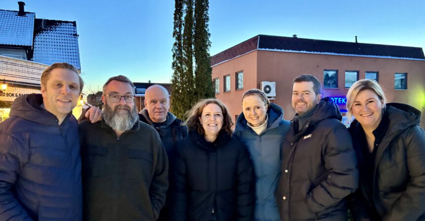 Gruppebilde av åtte personer smilende utendørs foran bygninger i klart vær.