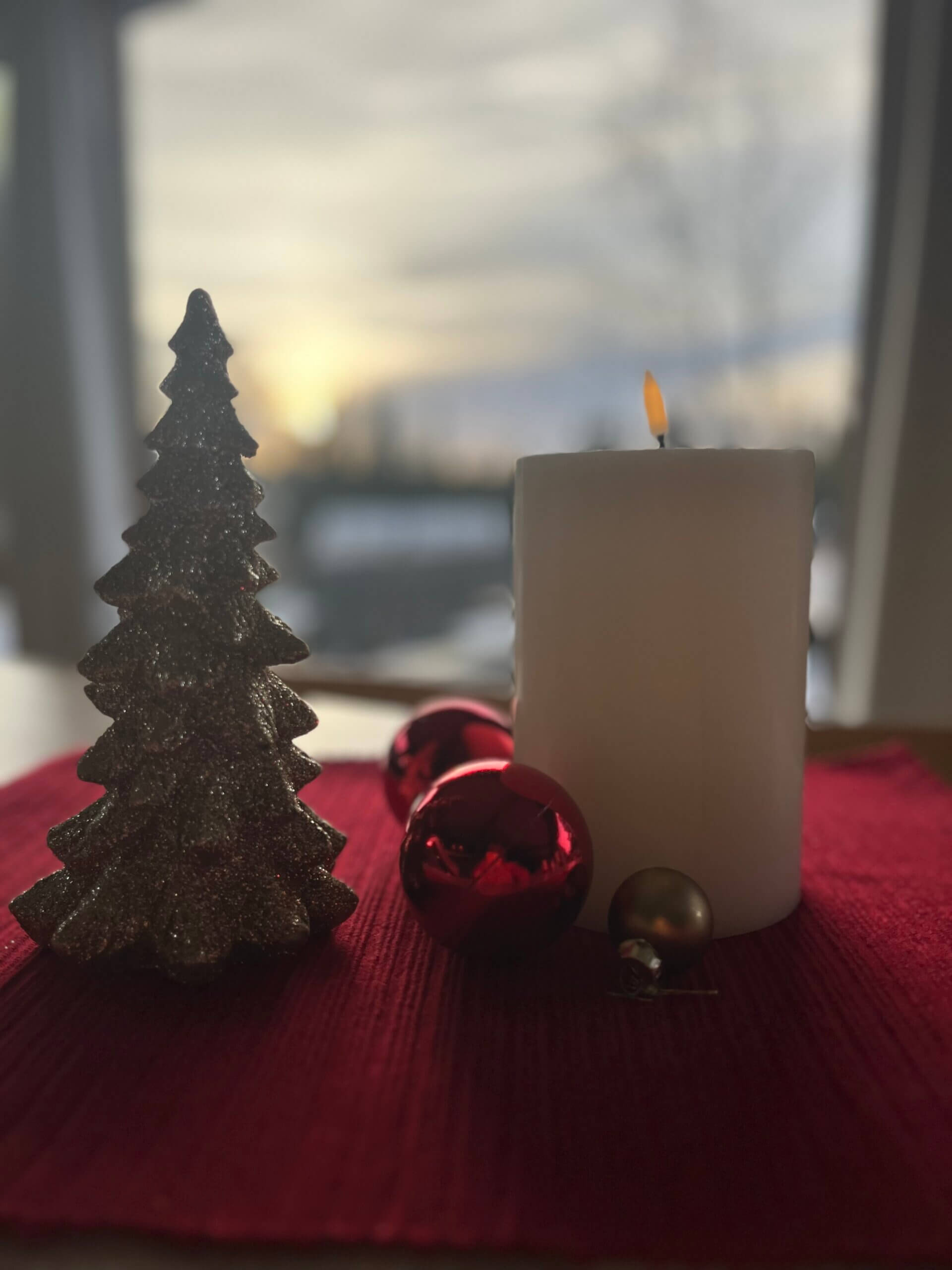 Julestemning med lys, juletrefigur, og røde julekuler på en rød duk. Vakkert vinterlys i bakgrunnen.