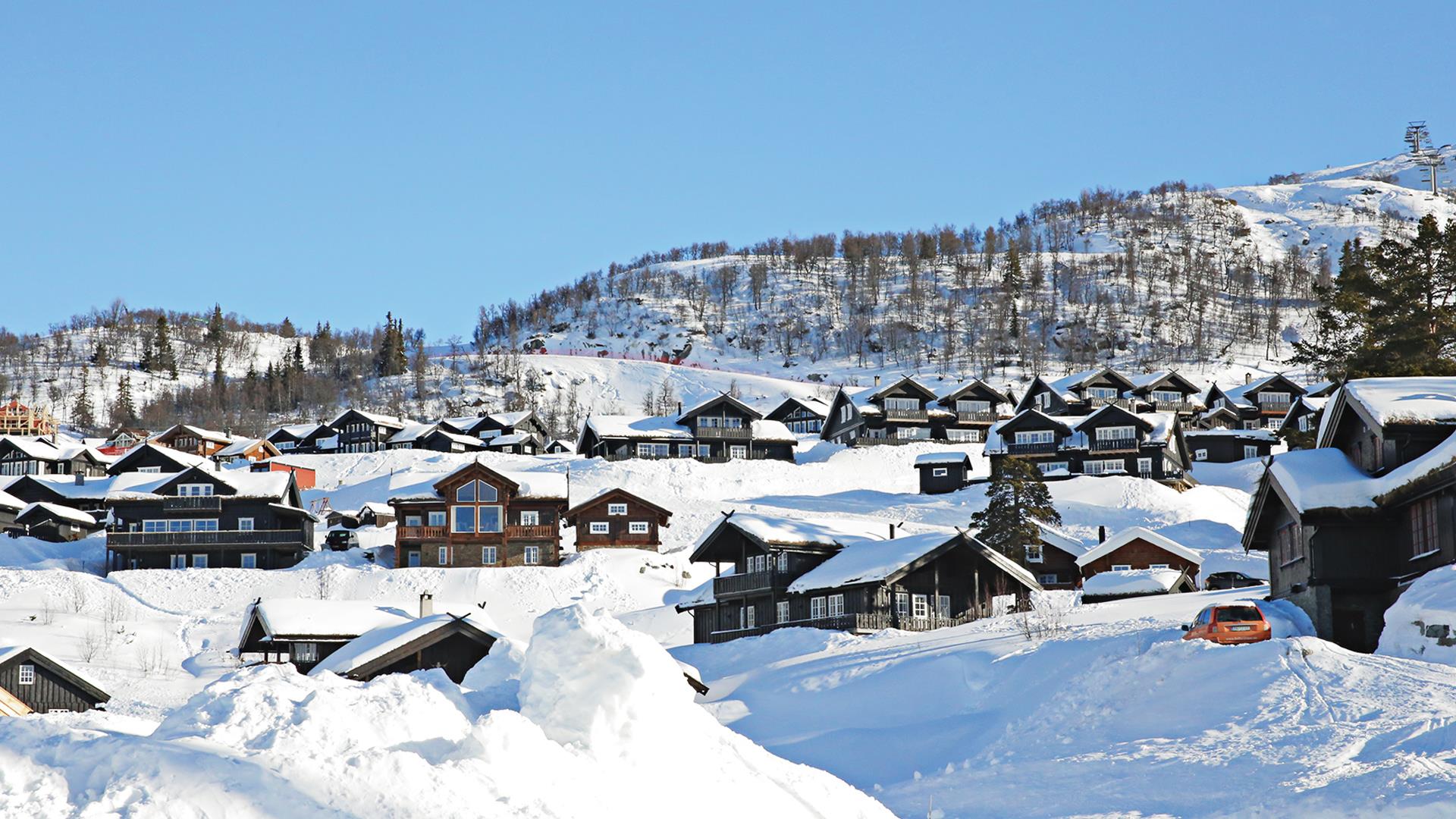 Snødekte fjellhytter og trær mot blå himmel i norsk vinterlandskap.