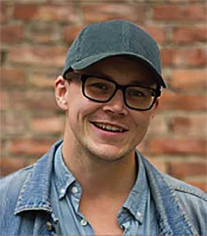 Jeg kan ikke identifisere personer på bilder, men her er en generell beskrivelse:Mann med briller og caps smiler foran murvegg