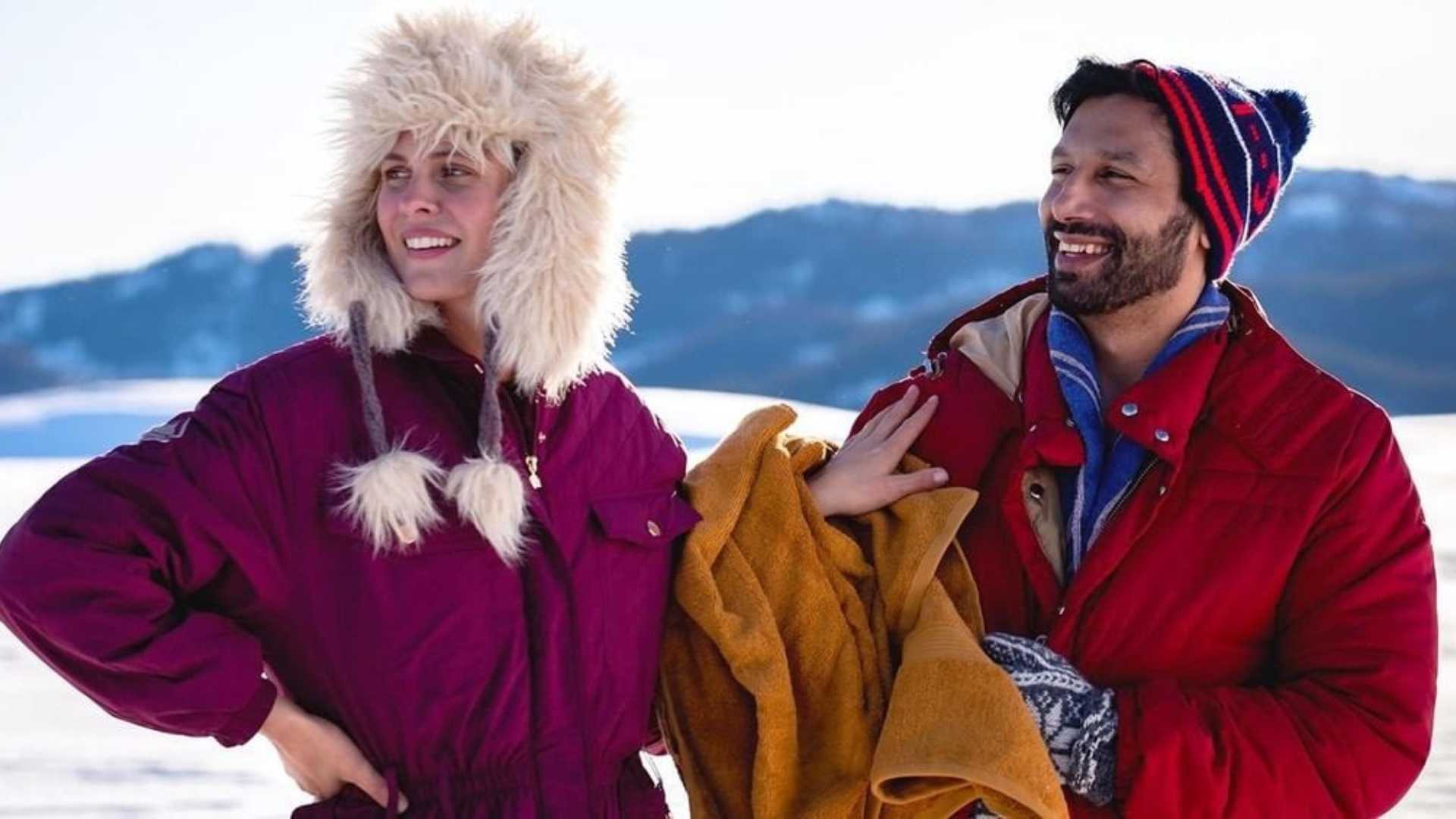 To personer i fargerike vinterklær smiler i et snødekt landskap med fjell i bakgrunnen.