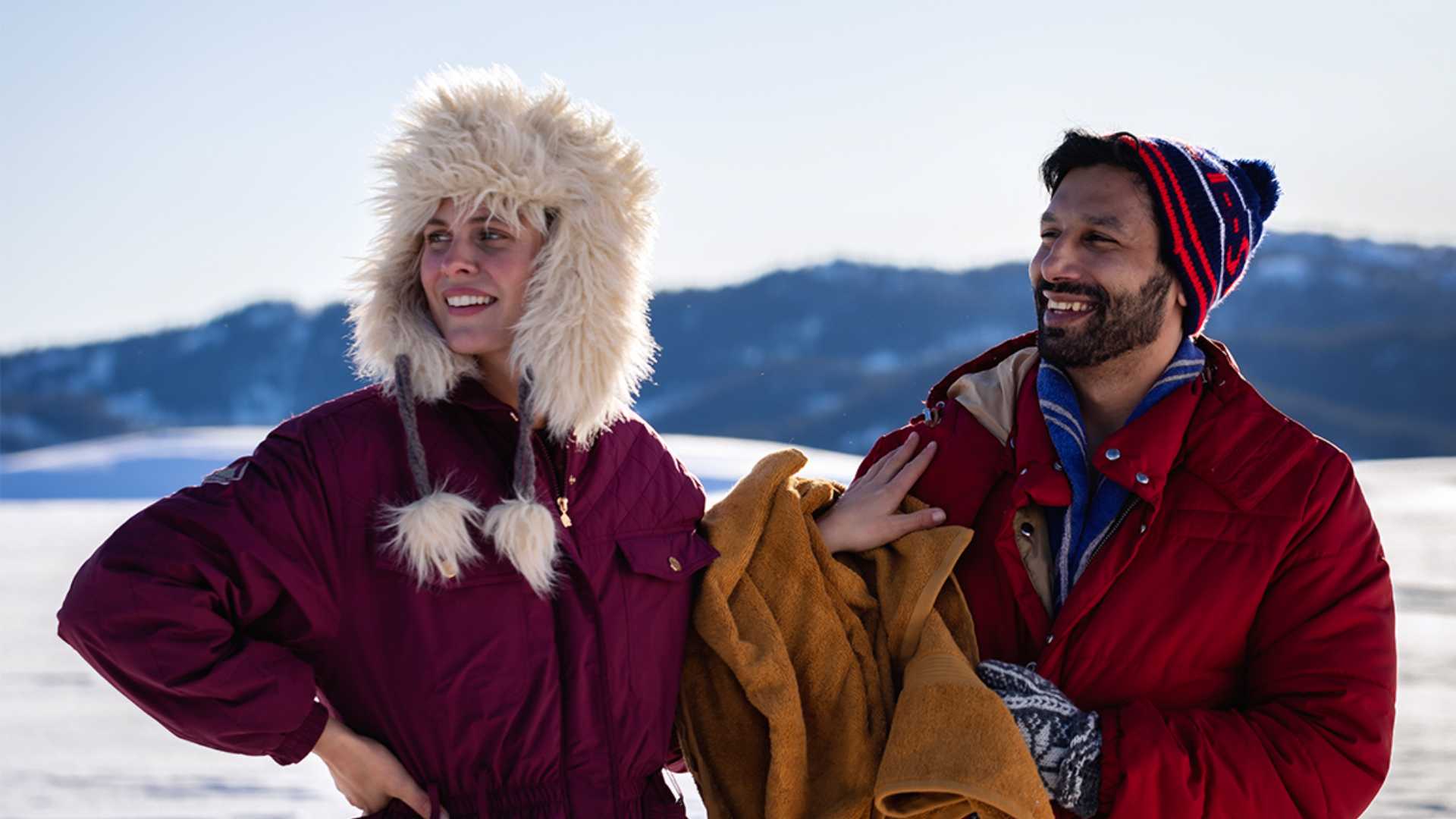 To personer i vinterklær smiler i snødekt landskap, iført lue og jakke. Bakgrunn med fjell og blå himmel.