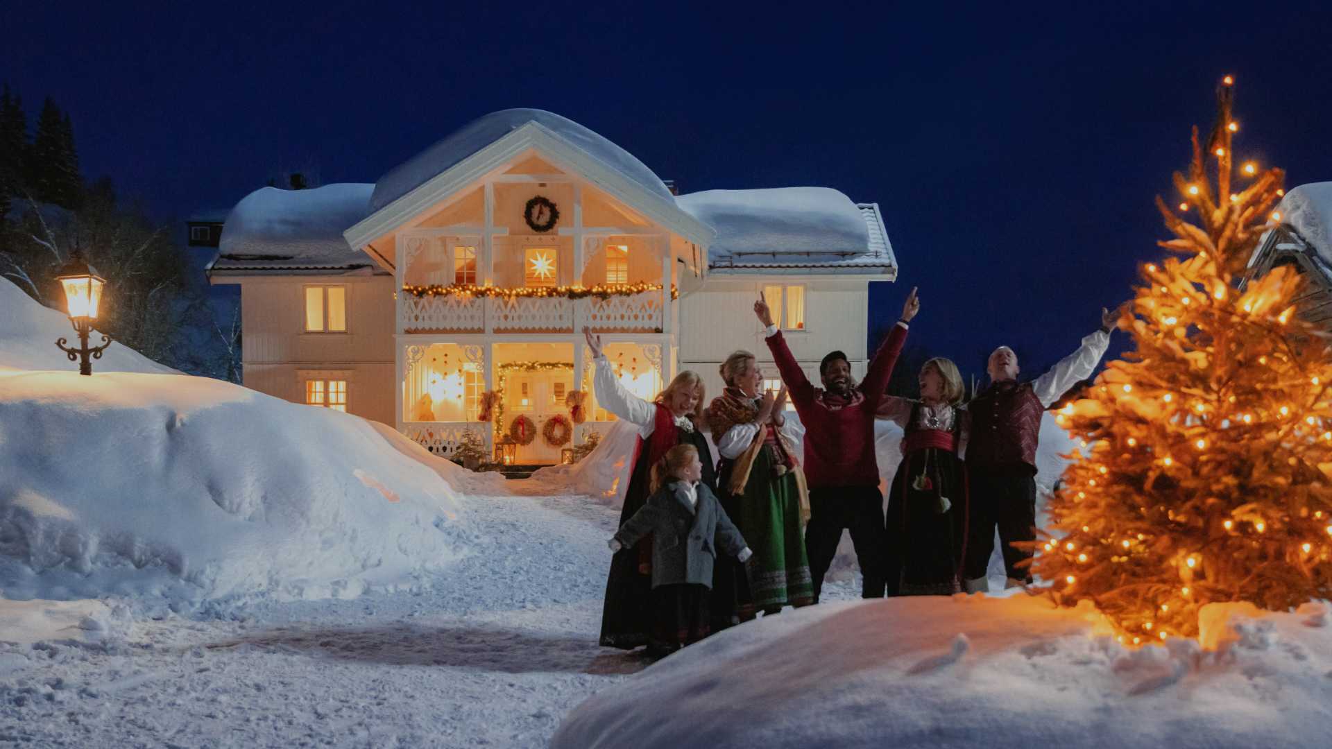 Feststemt familie i bunad foran et julepyntet hus og tre i vinterlandskap. Stemningsfull norsk julefeiring.