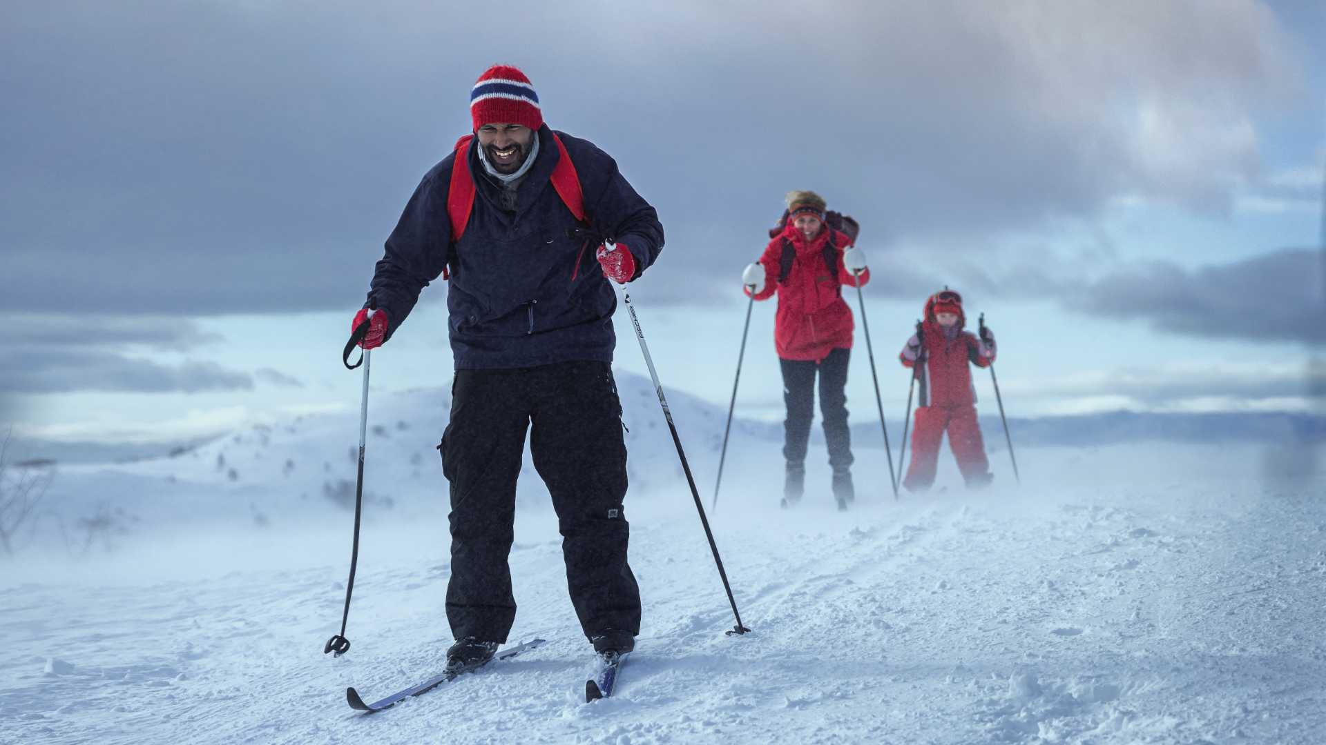 Tre personer går på ski i snødekt landskap under overskyet himmel.