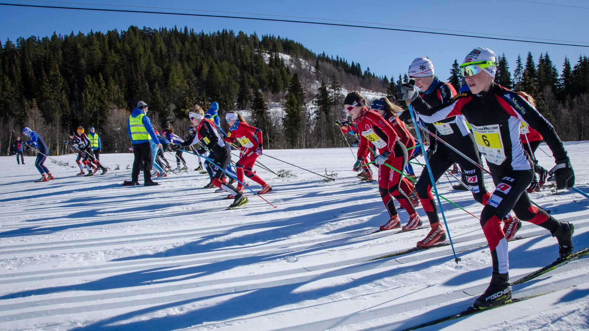 Langrennsløpere i startfasen av et skirenn i solfylt vinterlandskap med snø og skog.