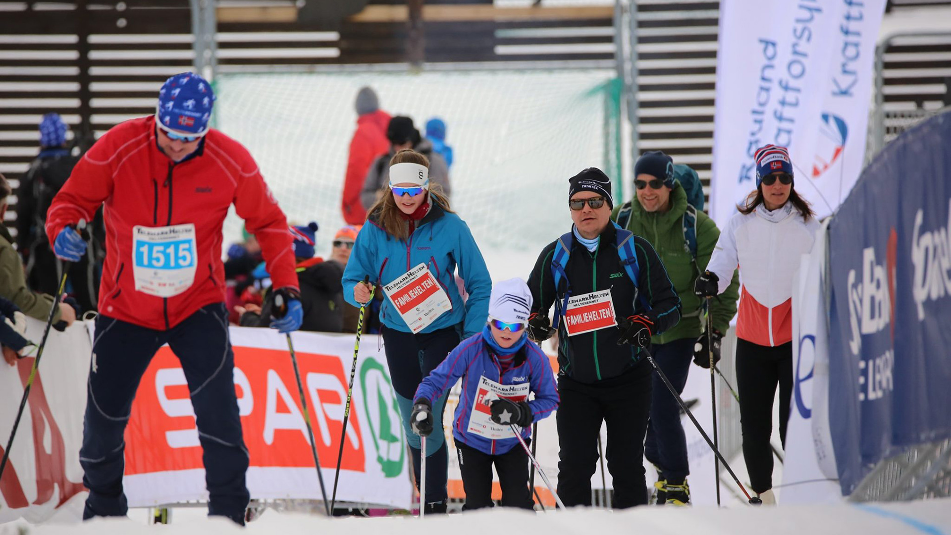 Familie på skirenn i Norge med startnumre og skiklær, mennesker i bakgrunnen og sponsorbannere.