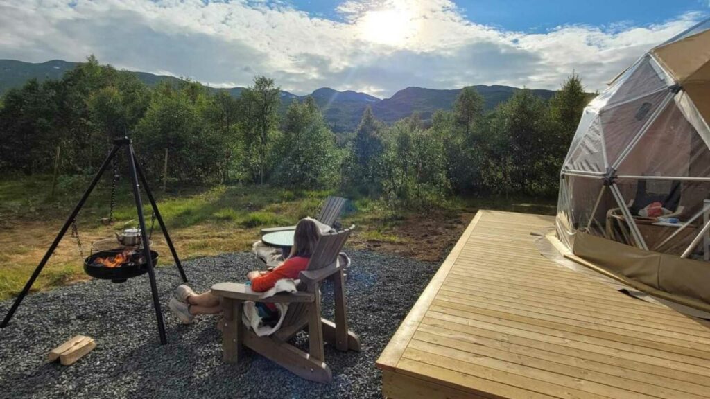 Person slapper av i stol ute ved telt, med ildsted og skog. Solen skinner over fjellene i bakgrunnen.