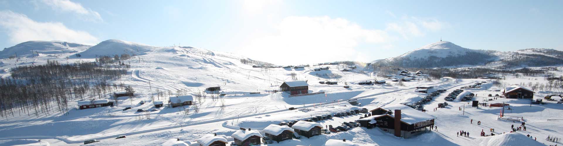 Snødekt skisted med fjell og hytter under klar blå himmel, skikjørere i bakgrunnen. Vinterlandskap i Norge.