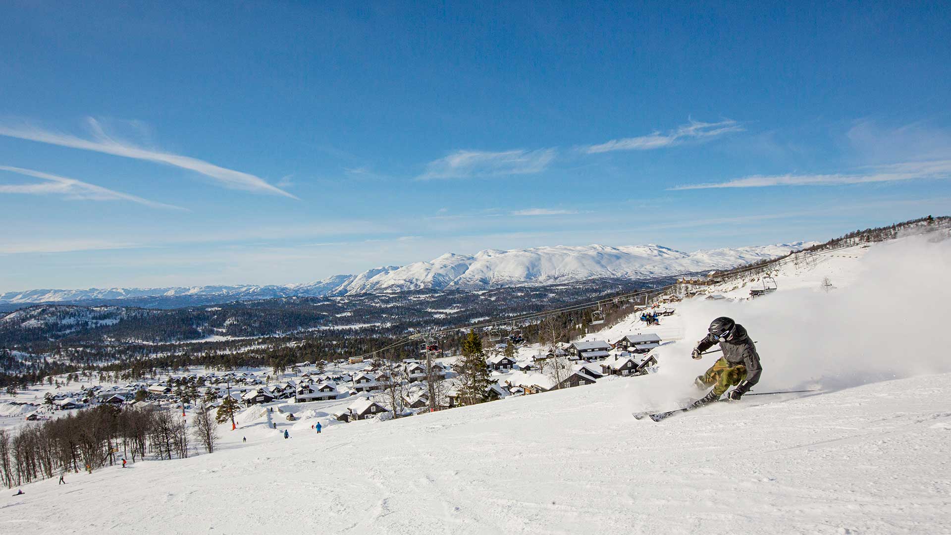 To personer i skiheis over snødekte fjelllandskap, klar himmel i bakgrunnen.