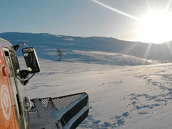 Tråkkemaskin i vinterlandskap med soloppgang over snødekte fjell.