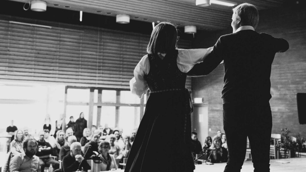 To personer danser på scenen foran et publikum i en sal, set bakfra.