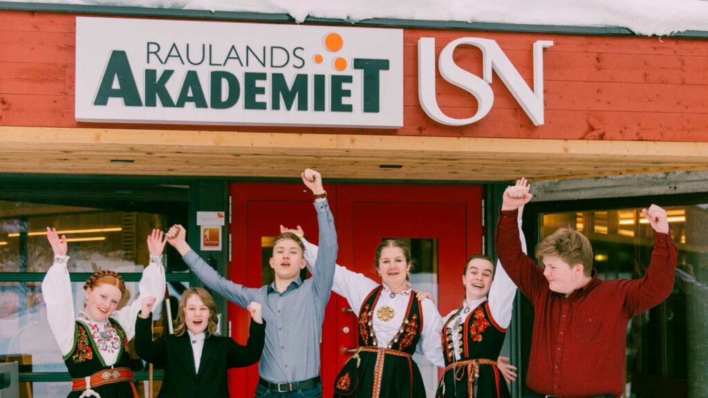 Gruppe mennesker foran skiltet til Raulandsakademiet, kledd i bunad, jubler med armene hevet.