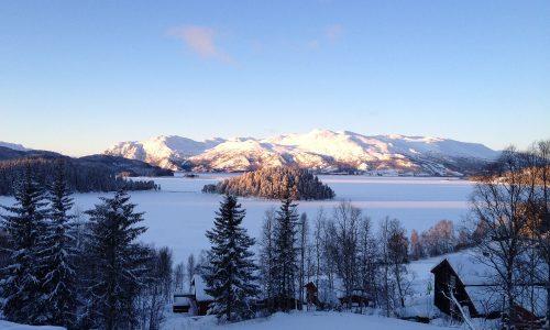 Snødekte fjell og frossen innsjø i solskinn, omgitt av grantrær og hytter i en vakker vinterlandskap i Norge.