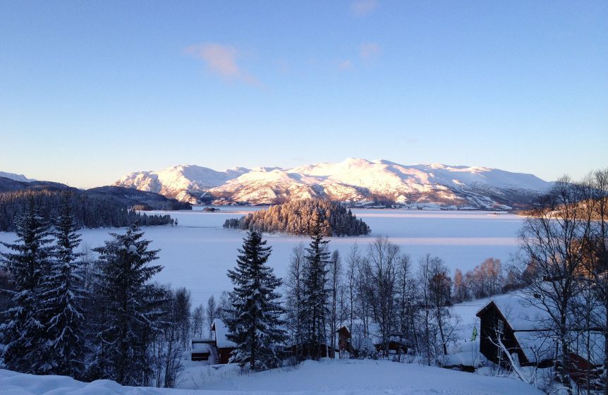 Snødekte fjell og frossen innsjø i solskinn, omgitt av grantrær og hytter i en vakker vinterlandskap i Norge.