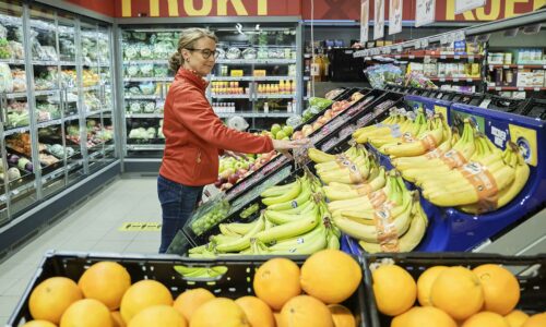 Kvinne velger frukt i supermarked, står ved banan- og sitronseksjonen. Ferskvarer i bakgrunnen.