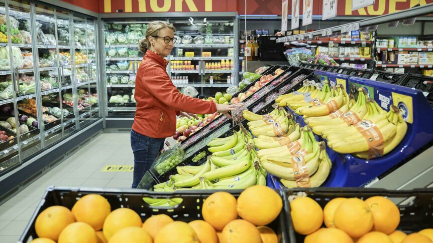 Kvinne velger frukt i supermarked, står ved banan- og sitronseksjonen. Ferskvarer i bakgrunnen.