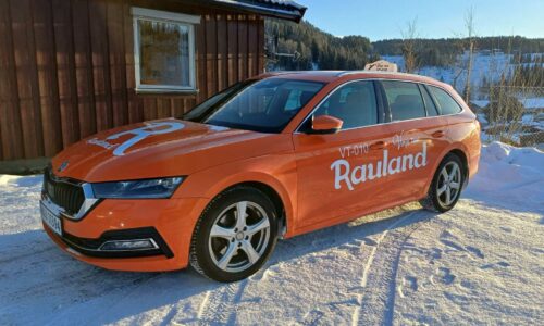 Oransje taxi med Rauland parkert i snødekt område foran en trebygning.