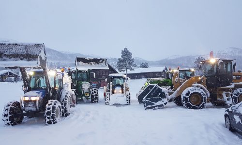 Traktorer rydder snø ved gårdshus i vinterlandskap, fjell i bakgrunnen.