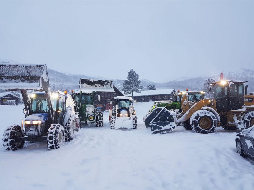 Traktorer rydder snø ved gårdshus i vinterlandskap, fjell i bakgrunnen.