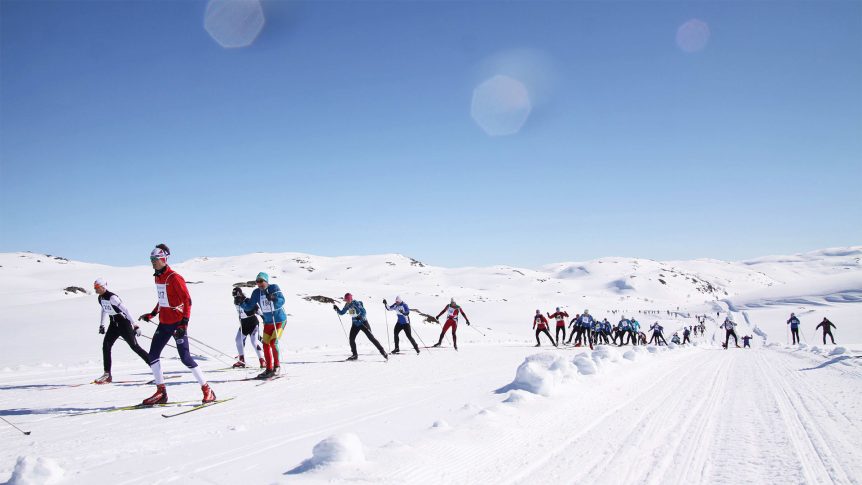 Deltakere i langrennsløp går på ski i snødekt terreng under klar blå himmel.