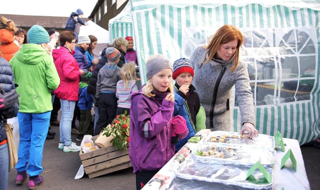 Gruppe mennesker ved et markedstelt smaker matprøver, mens barn venter spent.