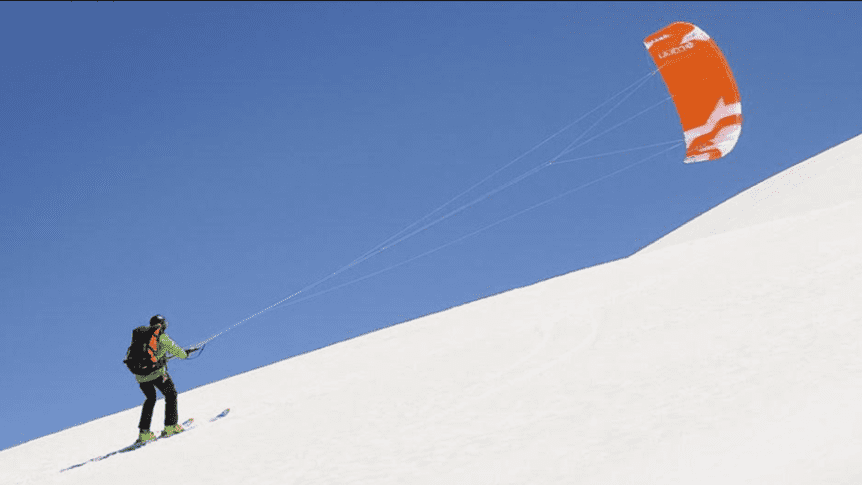 Person på ski kiter opp en snødekket bakke under blå himmel. Skikjøring med kite i vinterlandskap.