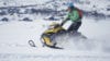Mann kjører snøscooter i snødekt landskap med fjellandsby i bakgrunnen. Vinteraktiviteter i Norge.