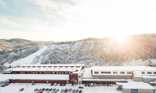 Vinterutsikt over et snødekket fjellandskap med en større bygning og parkerte biler i forgrunnen, sollys bak.