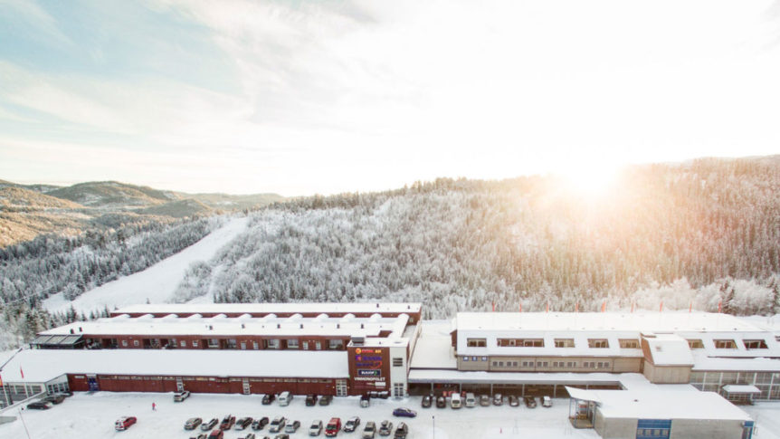 Vinterutsikt over et snødekket fjellandskap med en større bygning og parkerte biler i forgrunnen, sollys bak.