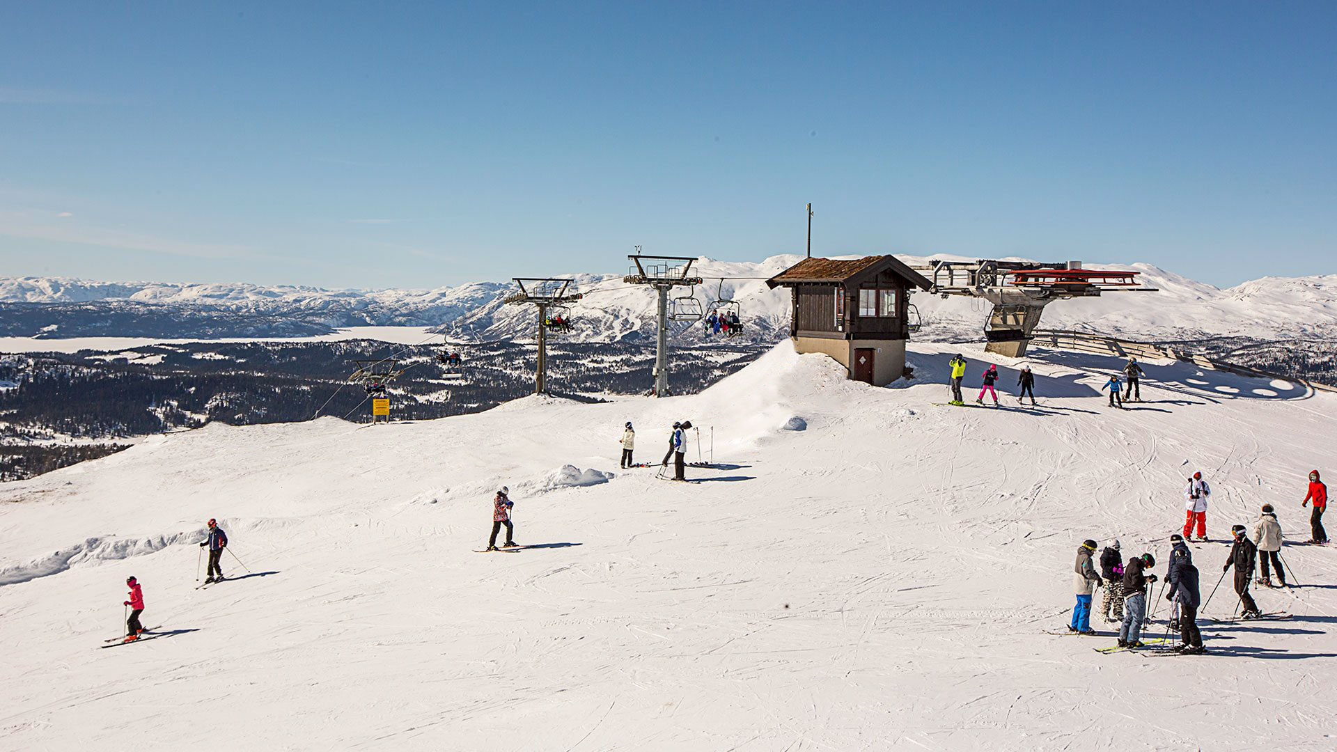 Skiløpere i et vintersportssted med fjell og skiheis i bakgrunnen på en solfylt dag.