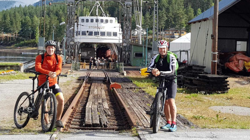 To syklister ved siden av en gammel ferge på en skinnegang omringet av trær og bygninger.