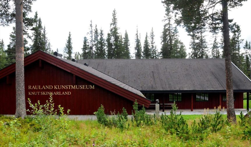 Rød trebygning av Rauland kunstmuseum omgitt av furutrær og gress.