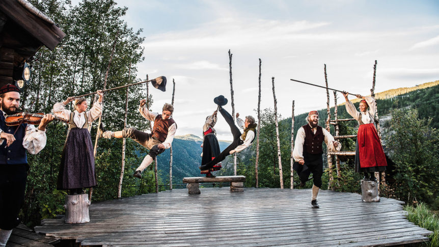 Tradisjonell norsk dans på utendørsscene, folk i bunader med fjellandskap i bakgrunnen.