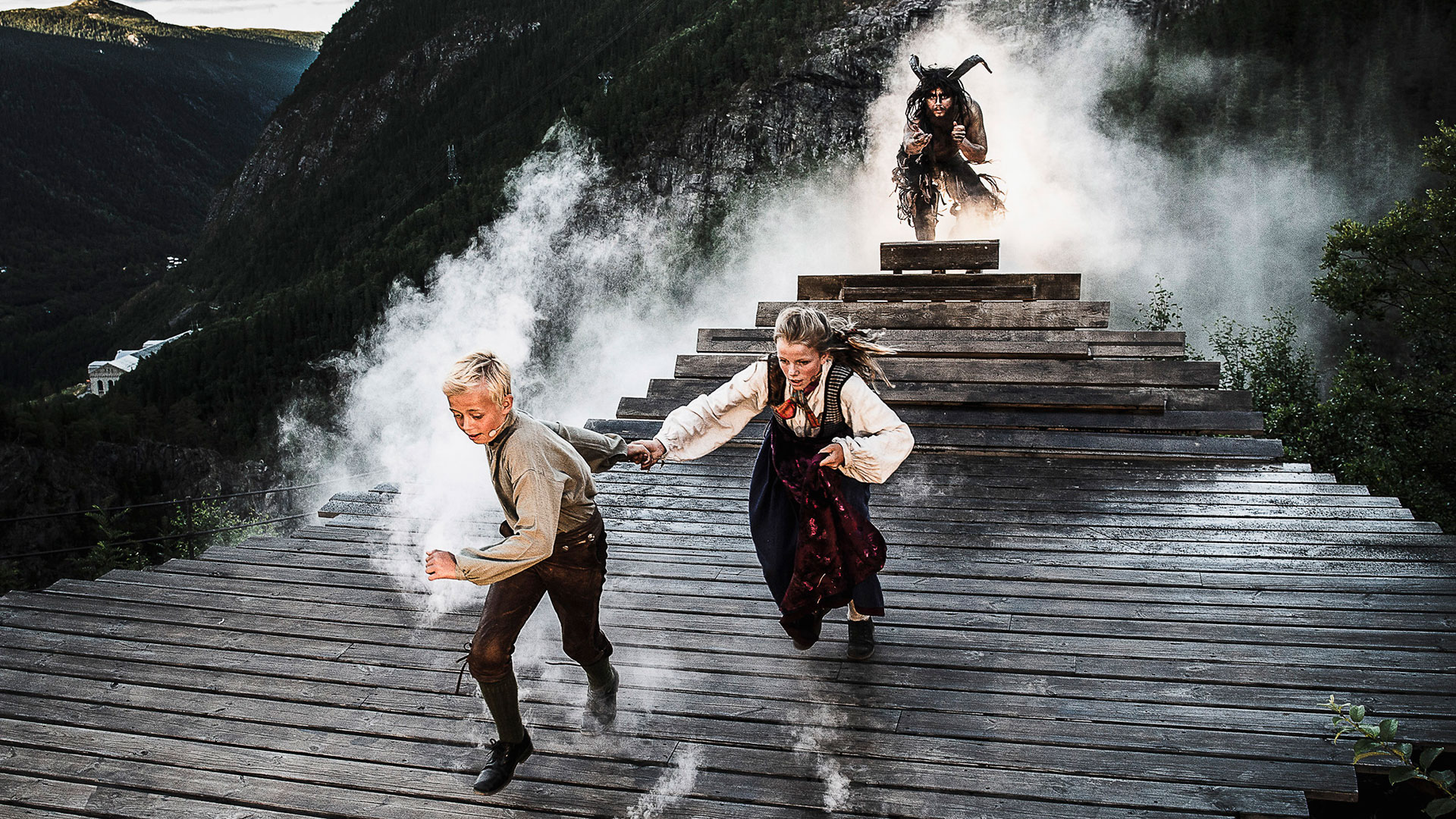 Barn løper på treplattform med mystisk skikkelse i røyken bak dem, omgitt av fjellandskap.