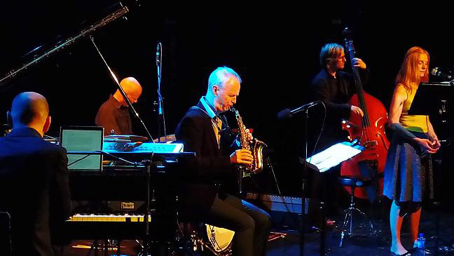 Jazzband opptrer på scenen med piano, saksofon, kontrabass og vokalist i lyseblått lys.