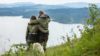 Par i turklær nyter utsikten over innsjøen fra en høyde i skogen, med en hund ved siden av.