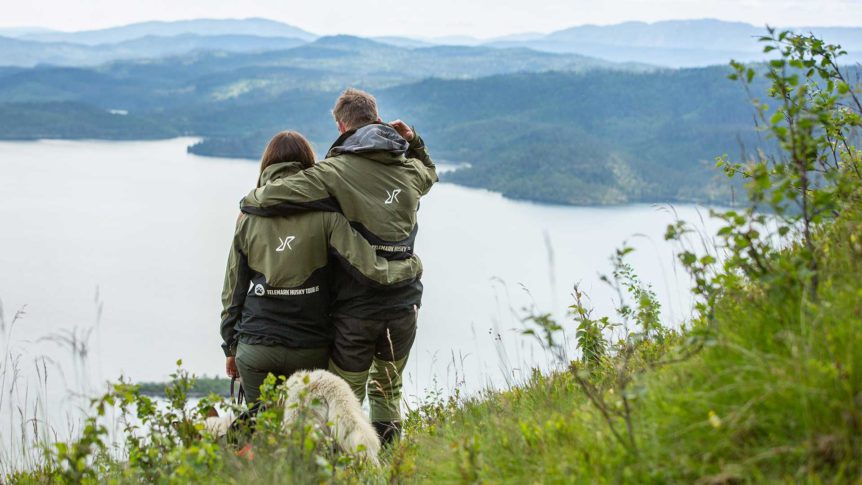 Par i turklær nyter utsikten over innsjøen fra en høyde i skogen, med en hund ved siden av.