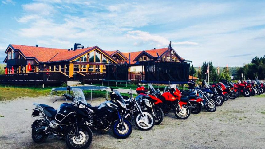 Rader med motorsykler parkert foran en stor, trebygning med rød takstein under en klar blå himmel.