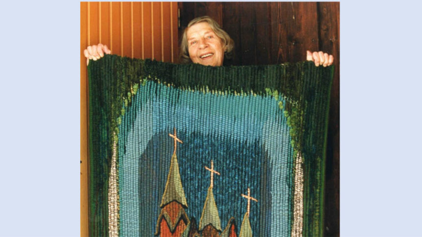 Kvinne smilende bak fargerikt teppe med kirketårn-design, mot en trevegg.