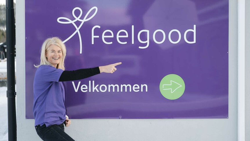 Kvinne i lilla t-skjorte peker på et skilt med teksten Feelgood Velkommen og en grønn pil, ute i snø.