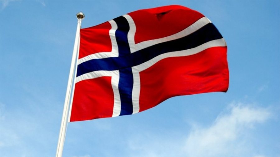 Norsk flagg vaier i vinden mot blå himmel, med rødt bakgrunn og blått kryss med hvit kant.